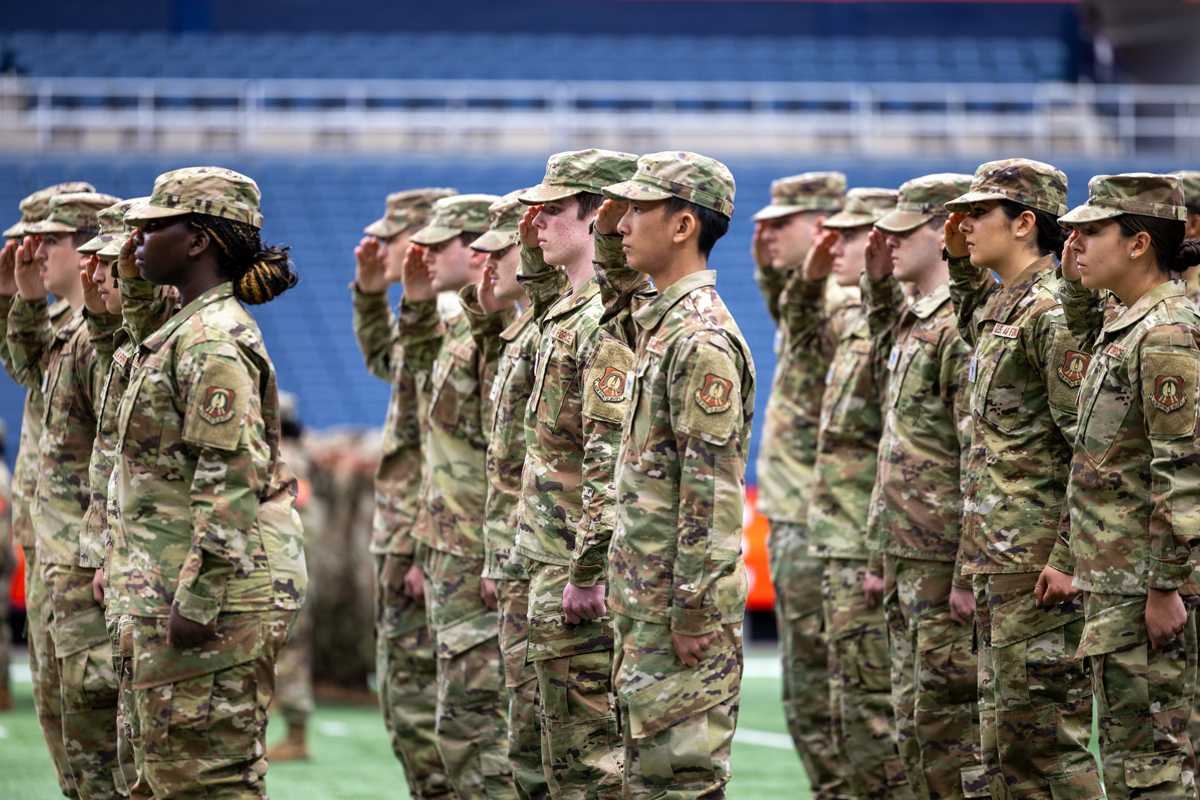 The image shows a group of soldiers in camouflage uniforms standing in formation, saluting. The soldiers are arranged in rows, facing forward. The background shows an indoor setting with blue seating and a railing above the soldiers.
