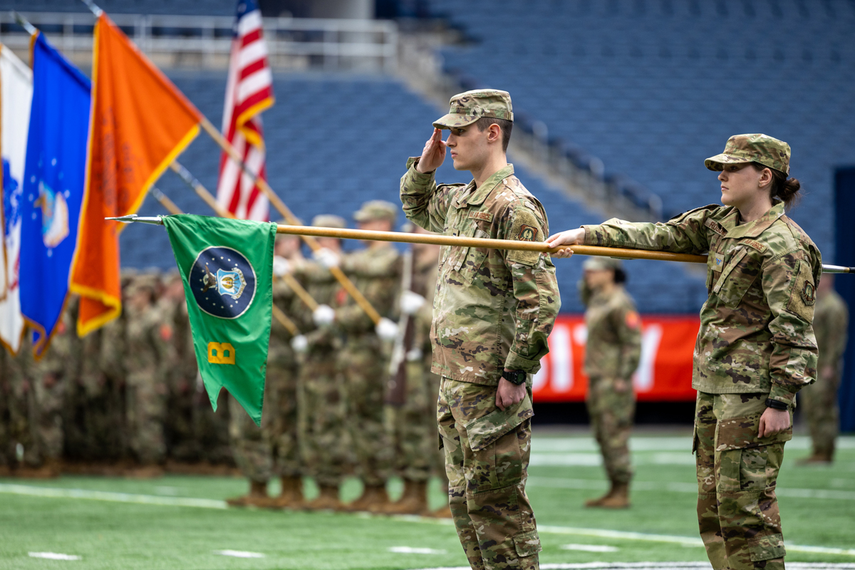 The image shows a military ceremony taking place on a field. Two soldiers in camouflage uniforms are standing in the foreground; one is saluting while the other holds a flagpole with a green flag that has an emblem and yellow symbols on it. In the background, there are more soldiers standing in formation, holding various flags including an American flag and other colorful flags. The setting appears to be indoors, possibly in a stadium or large arena with blue seating visible.