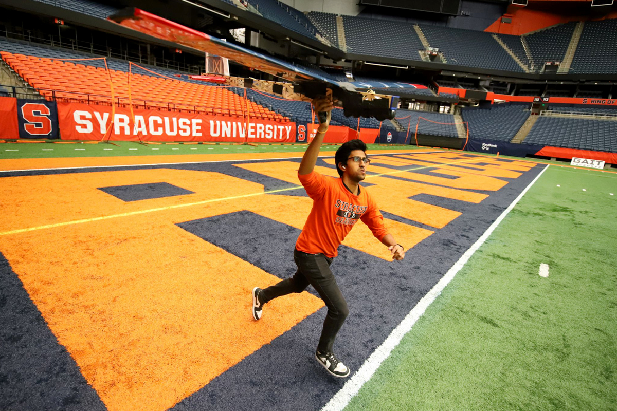 A person wearing an orange shirt and black pants is running on a football field with an airplane. The field has large orange letters spelling out "SYRACUSE" on the blue end zone area. In the background, there are empty stadium seats and a banner that reads "SYRACUSE UNIVERSITY."