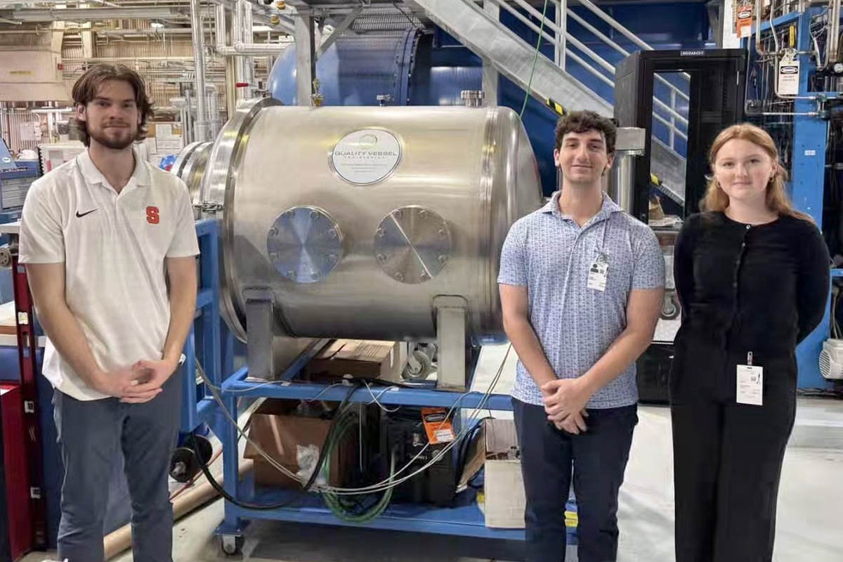 Three people are standing in front of a large metallic cylindrical machine with two circular windows. The machine is mounted on a blue frame with wheels and has various cables and equipment around it. The background shows an industrial setting with stairs, pipes, and other machinery.