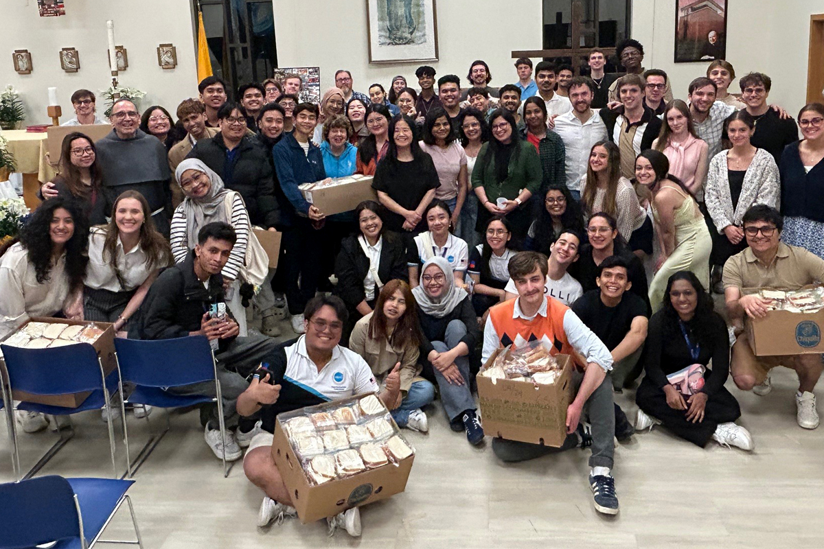 The image shows a large group of people gathered in a room, posing for a photo. Some individuals are holding boxes filled with sandwiches. The room has chairs arranged in rows, and there are various decorations on the walls, including framed pictures and plants.