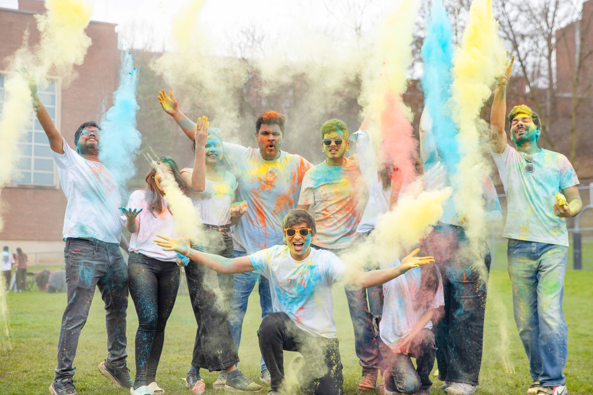 Students throw brightly colored powder up in the air while smiling and celebrating the Holi Festival.