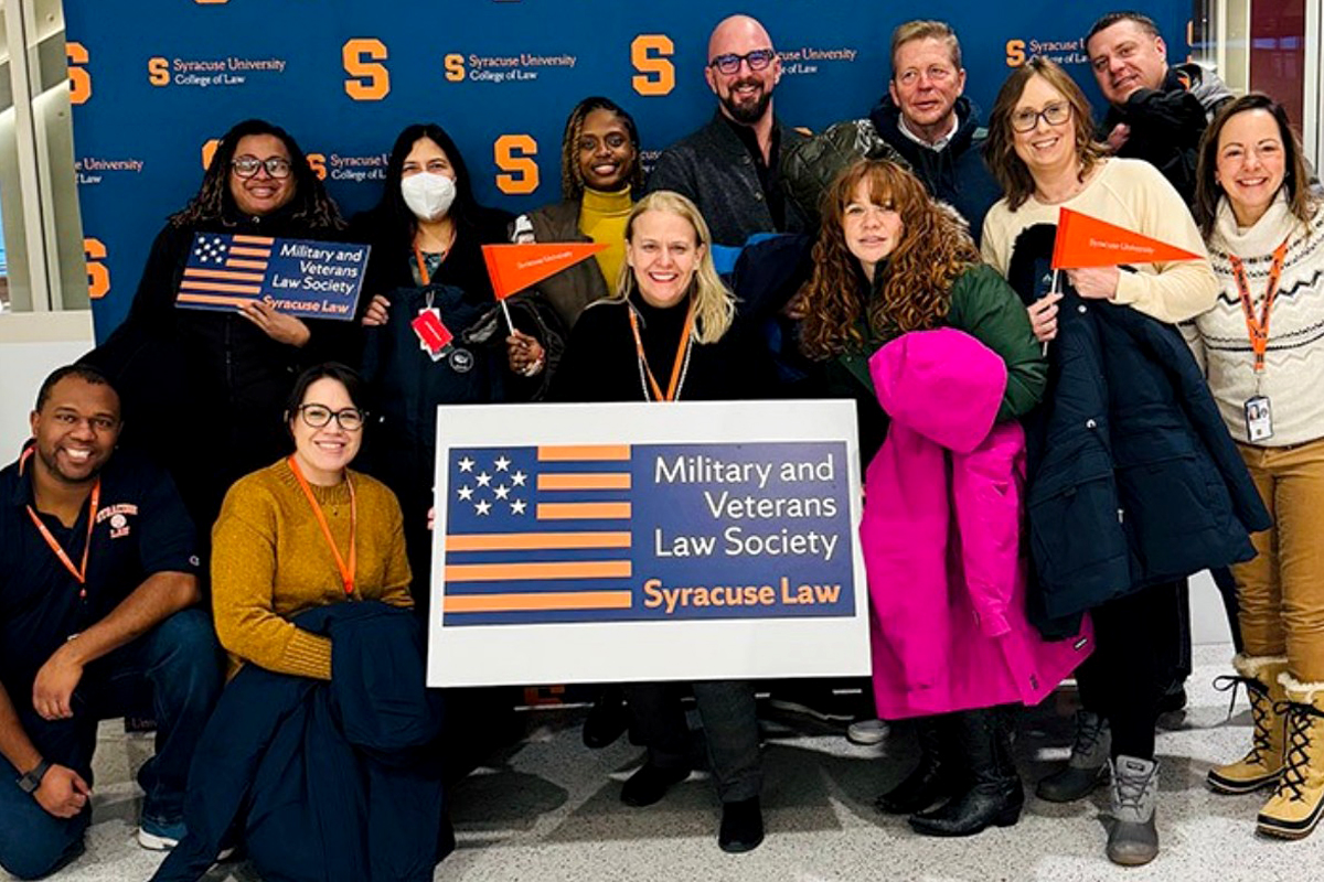 The image shows a group of people standing in front of a backdrop with the Syracuse University College of Law logo. They are holding signs and banners that read "Military and Veterans Law Society" and "Syracuse Law." Some individuals are wearing lanyards, and one person is holding a pink coat.