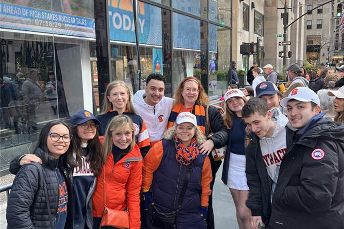 The image shows a group of people standing outside a building with glass windows. The individuals are dressed in various orange, blue, and white clothing items, some of which have "Syracuse" written on them. The background shows a sign that reads "TODAY"