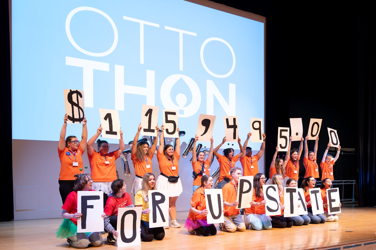 The image shows a group of people on a stage holding up large signs with numbers and letters. The signs collectively display the amount "$115,749.50" and the words "FOR UPSTATE." The background features a large screen with the text "OTTO THON."