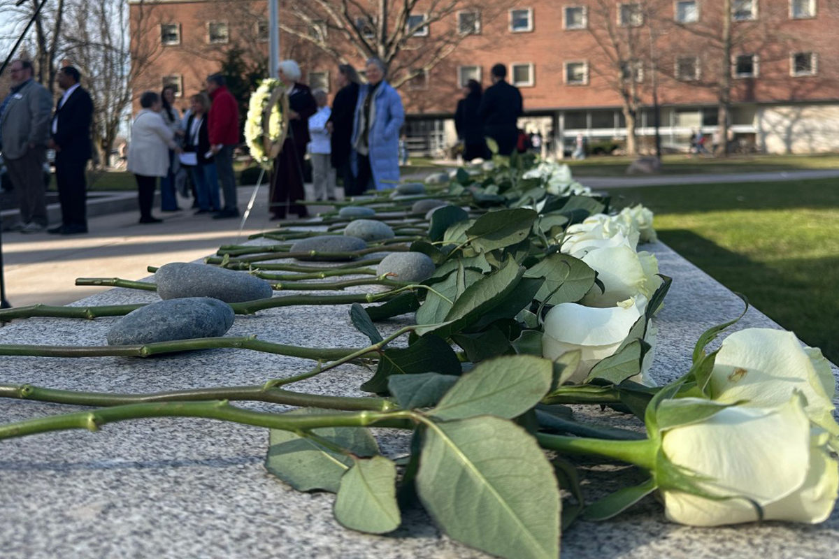 The image shows a memorial scene with white roses and stones placed on a stone surface. In the background, there are people gathered, some standing and some walking, in an outdoor setting with a building visible. The focus is on the flowers and stones in the foreground.