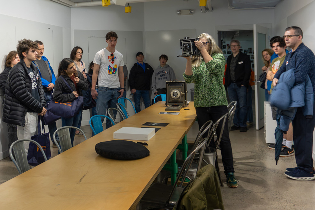 The image shows a group of people gathered in a room with white walls and various pieces of equipment. A person in the foreground is holding a large camera and appears to be taking a photograph. The group includes individuals standing around a long wooden table with metal chairs, some wearing jackets and casual clothing. There are books and other items on the table, including another camera setup. The room has overhead lighting and some posters or pictures on the walls in the background.