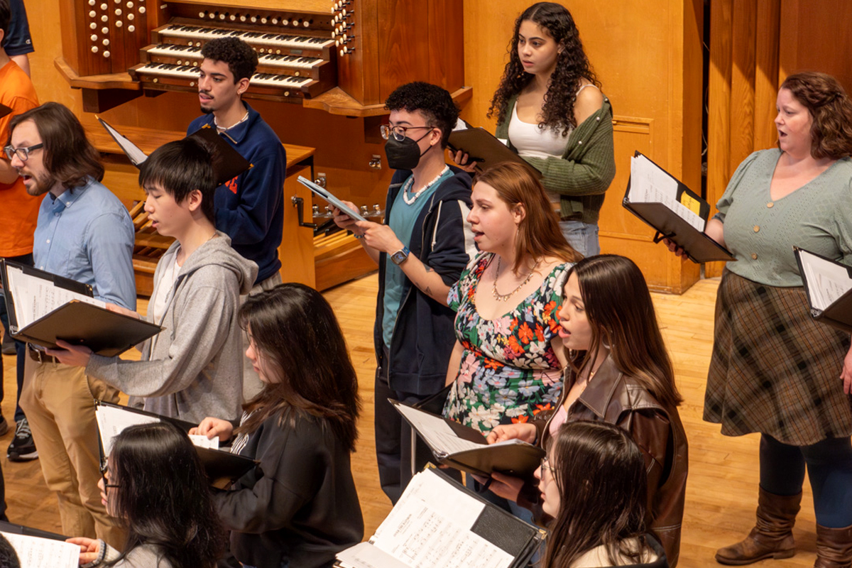 A group of people are standing in a room, holding sheet music and singing. The background features a large organ with multiple pipes and keyboards. The individuals appear to be participating in a choir rehearsal or performance.