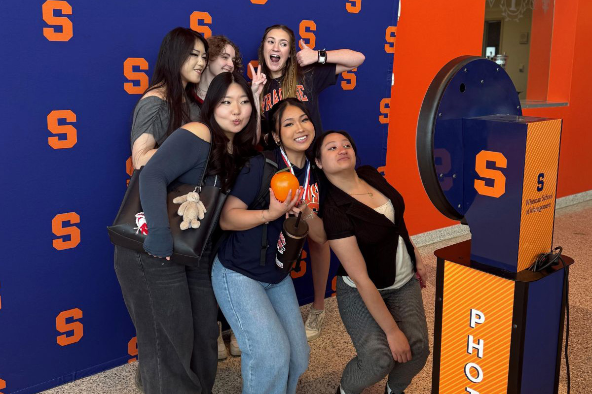 A group of people pose for a photo in front of a backdrop with multiple orange Block S's on a blue background. They are standing next to an orange and blue photo booth machine. One person is holding an orange balloon.