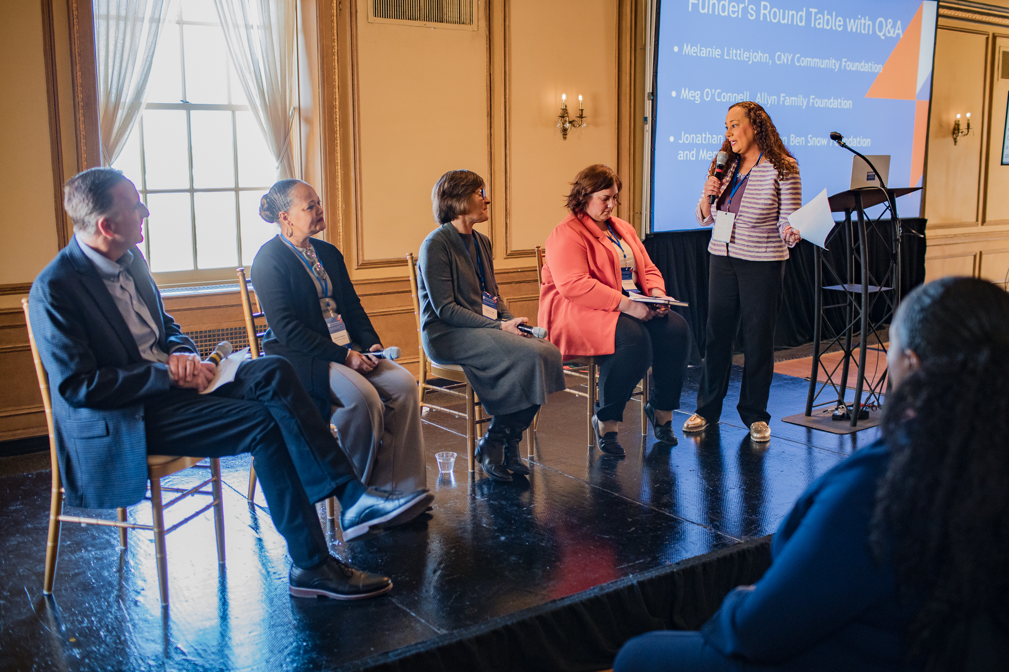 The image shows a group of five people seated on a stage in a panel discussion setup. Four individuals are sitting on chairs, while one person stands and speaks into a microphone. The background features a large screen displaying text that reads: "Funder's Round Table with Q&A," followed by the names "Melanie Littlejohn, DIR Community Foundation," "Meg O'Connell, Allyn Family Foundation," and "Jonathan Logan, Ben Snow Foundation." The room has elegant decor with chandeliers and large windows.