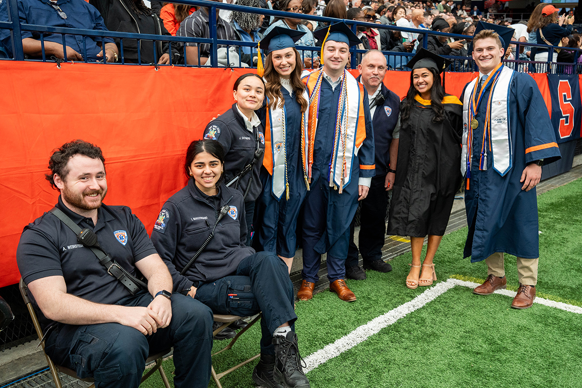 A group of people dressed in graduation attire and emergency responder uniforms, standing and sitting on chairs on a field. There is an orange banner behind them with a crowd of people seated in the background. The individuals are wearing caps, gowns, and stoles, indicating that they are graduates. Some individuals are also wearing uniforms with patches and badges.